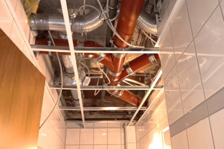 Rote Gussrohre der Abwasserinstallation oberhalb einer geöffneten abgehängten Decke in einem gefliesten Badezimmer.