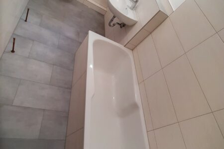 Modernisiertes Badezimmer mit Badewanne, Wand-WC, Waschtisch und großformatigen Wand- und Bodenfliesen.