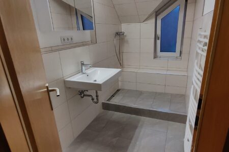 Modernisiertes Dachgeschossbad mit begehbarer Dusche, Waschtisch, Handtuchheizkörper und grauen Bodenfliesen.