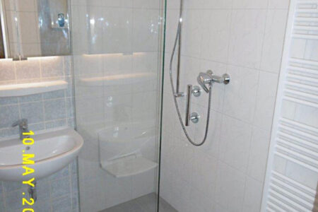 Modernisierte bodengleiche Dusche mit Glasabtrennung, Wandarmatur und Handbrause neben Waschbecken und Handtuchheizkörper.