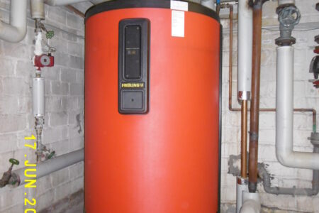 Großer roter Fröling Warmwasserspeicher mit angeschlossenen und gedämmten Rohrleitungen in einem Heizungsraum.