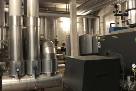 Große Heizungsanlage mit Heizkessel, Brenner und umfangreich gedämmten Rohrleitungen in einem gewerblichen Heizraum.