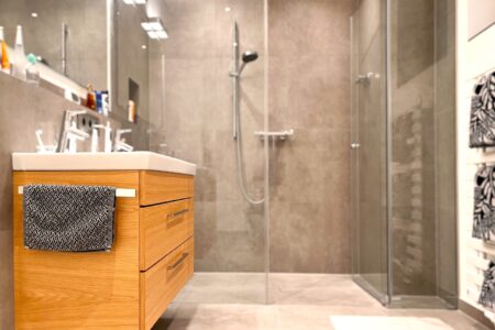 Modernes Badezimmer mit bodengleicher Glasdusche, großformatigen Fliesen und Waschtisch aus Holz.