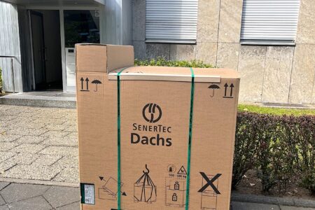 Verpacktes SenerTec Dachs Blockheizkraftwerk auf einer Holzpalette vor einem Gebäude.