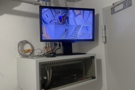 Monitor einer Videoüberwachungsanlage mit mehreren Kameraansichten über einem Netzwerkschrank.