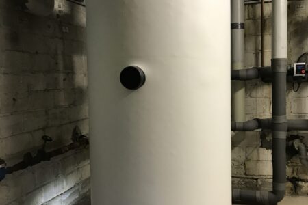 Großer weißer Warmwasserspeicher mit Temperaturanzeige und angeschlossenen gedämmten Rohrleitungen in einem Heizungsraum.